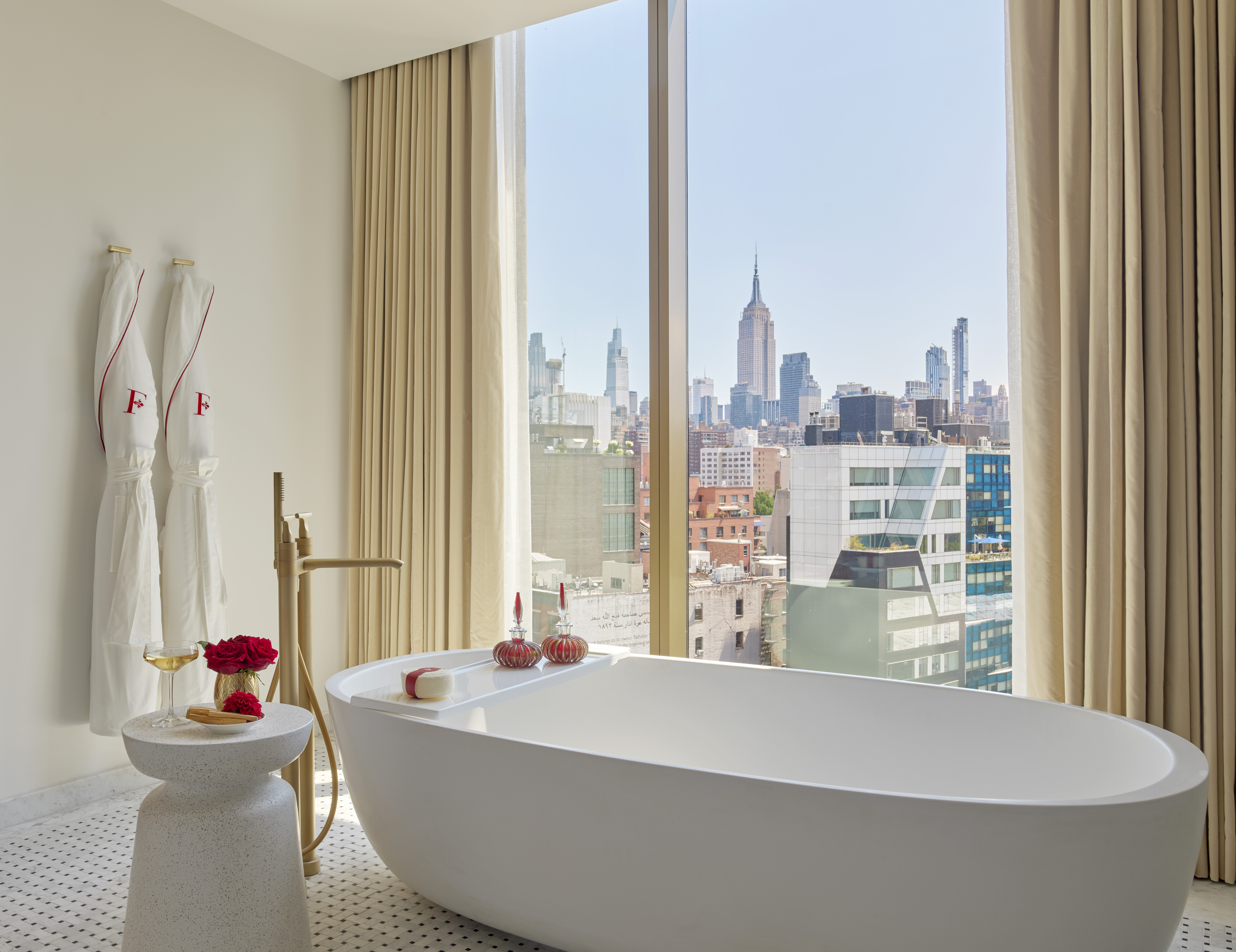A hotel bathroom with a large window overlooking the New York City skyline, including the Empire State Building.