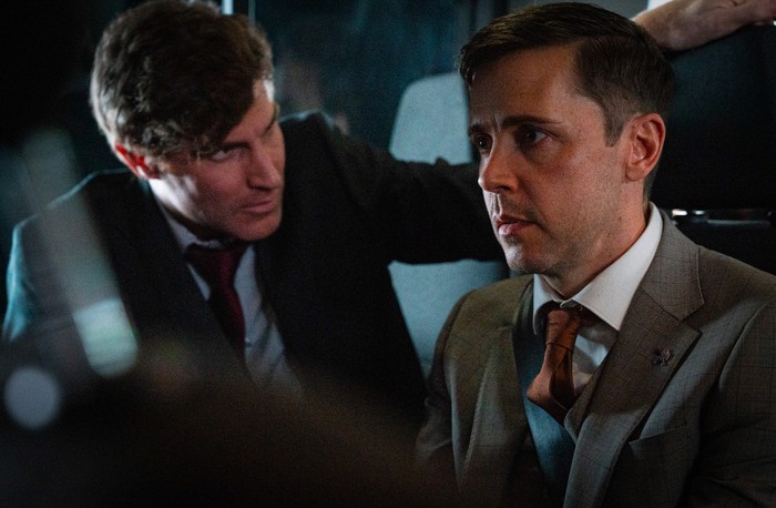 05_01_CORRIEDALE_THEO_TODD_01 Theo looks over Todd's shoulder in a scene from Corriedale. Both men are wearing suits.