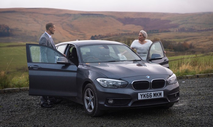 05_01_coro_carl_debbie_04 Debbie, in her wedding dress, stands opposite Carl, in a suit, as they are surrounded by hills and next to a car in a scene from Coronation Street