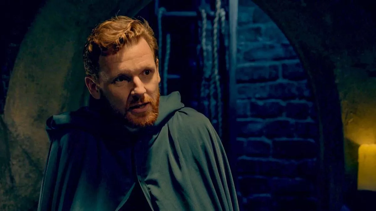 An individual with a beard, donning a green hooded cloak, stands in a dimly lit room with an intricate background.