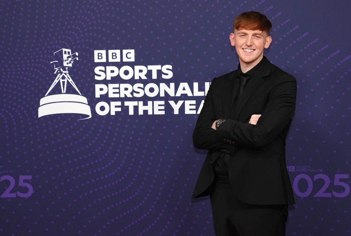An individual stands confidently on a stage, wearing a formal black suit, against a backdrop adorned with the BBC Sports Personality of the Year logo and branding. The person appears to be posing for an official photograph.