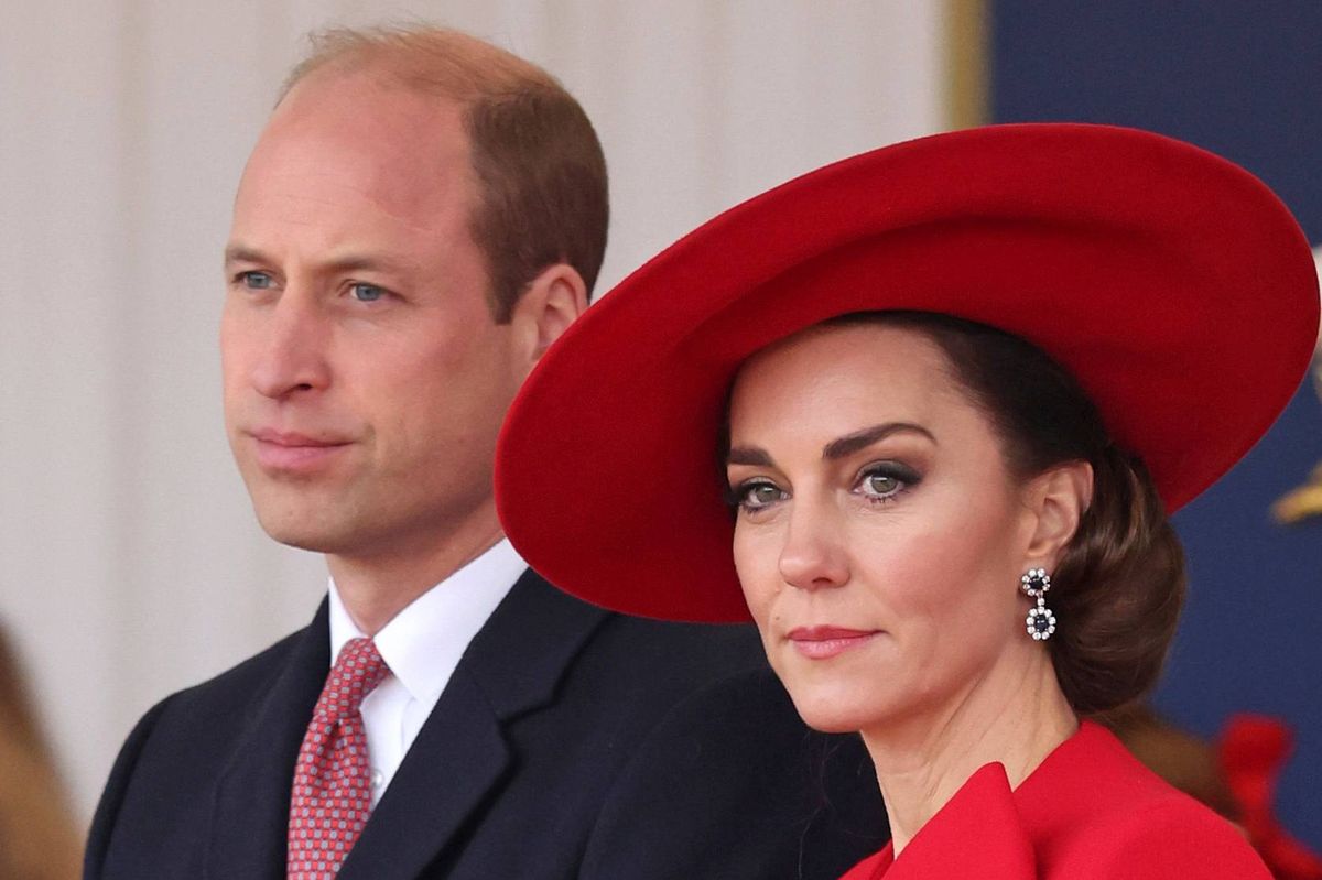 A formal image depicts a man and a woman standing side by side, likely attending an official event. The man is dressed in a black suit with a red tie, while the woman is wearing a red dress and a wide-brimmed red hat. Both individuals appear to be engaged in the proceedings, exuding a sense of elegance and formality.