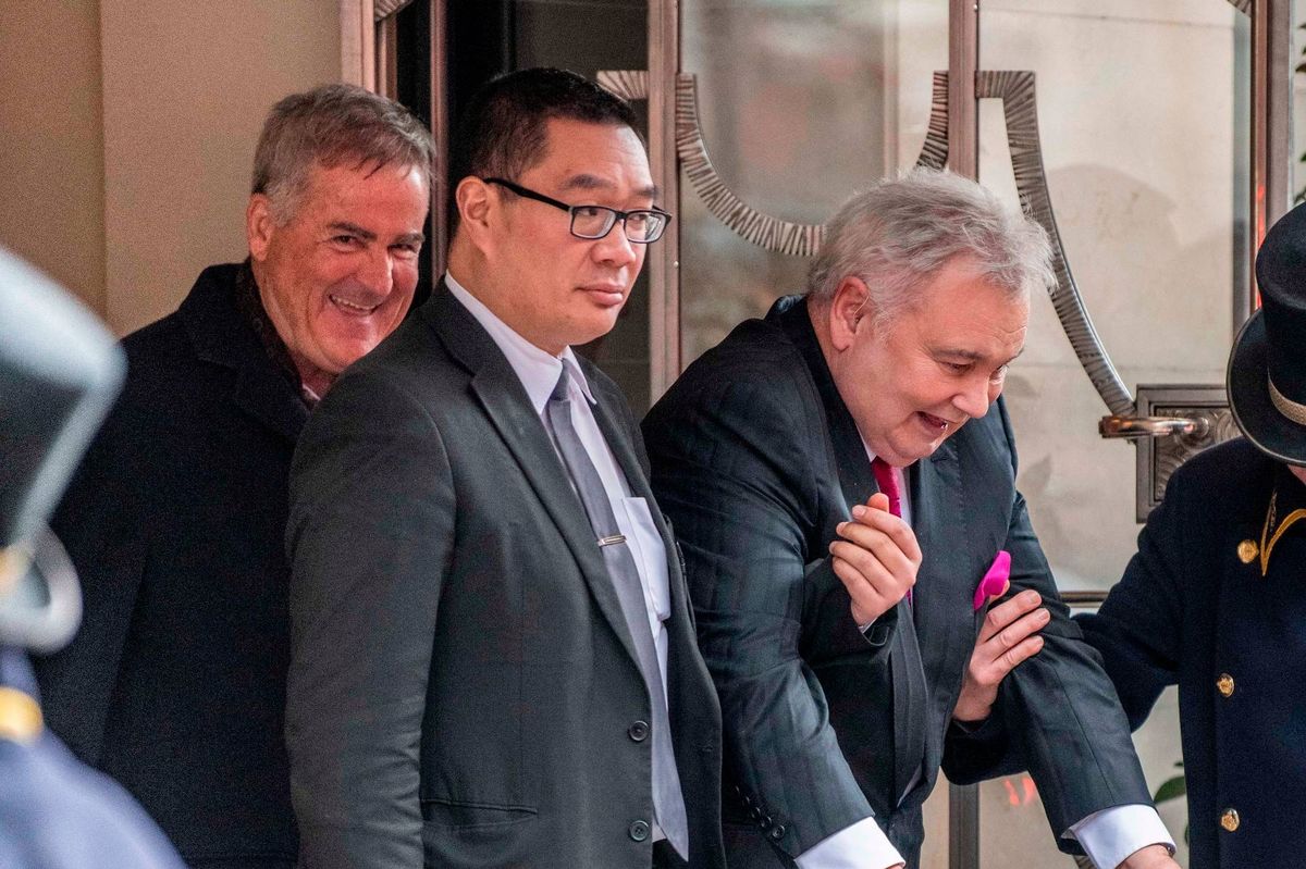 Three formally dressed individuals are standing together outside a building. One of them is wearing glasses and appears to be conversing with the person beside him. Behind them, a person in a blue uniform is partially visible, indicating the presence of security or law enforcement.