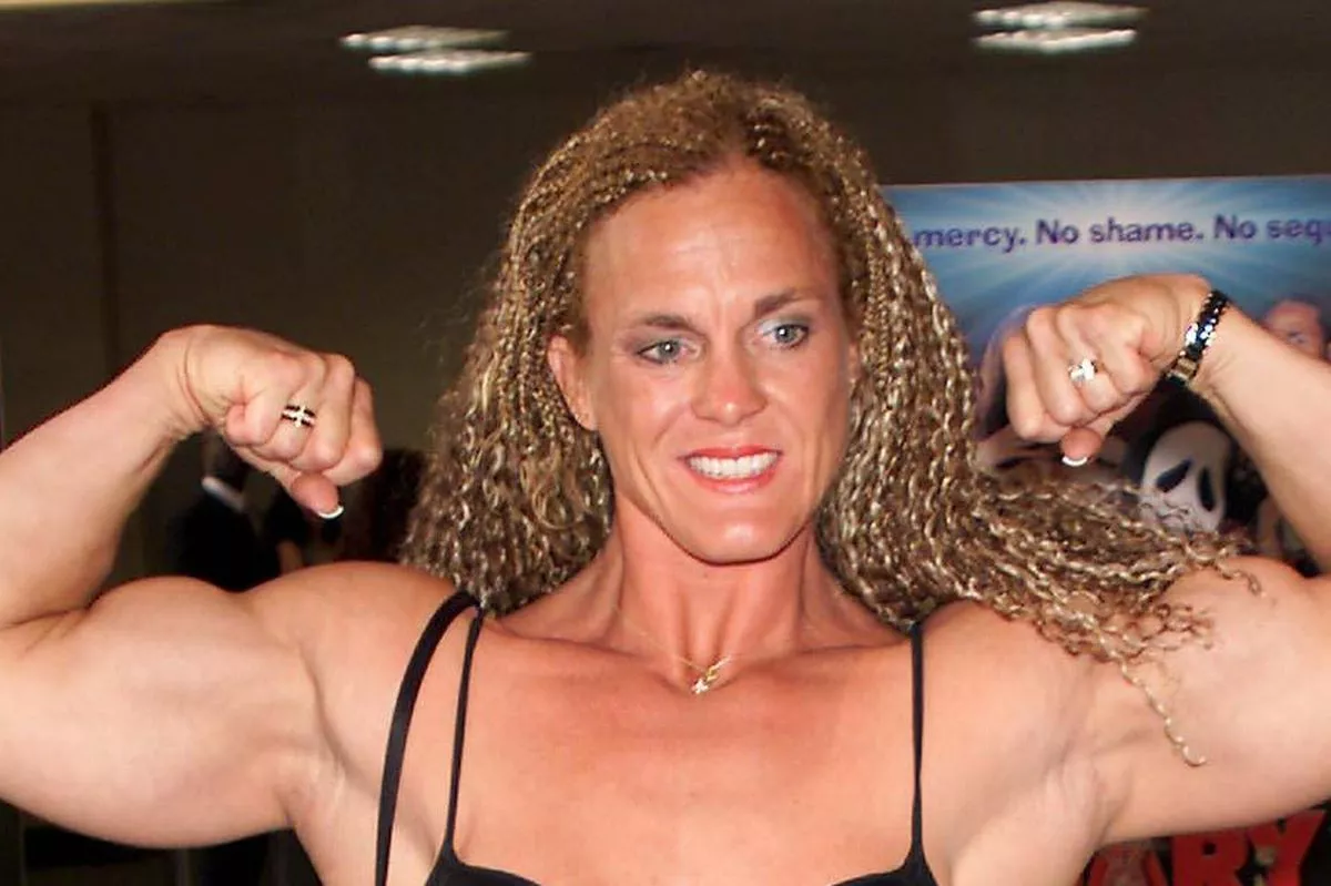 An adult female, dressed in a black cut-out dress, flexes her muscular arms in a room adorned with various posters in the background.