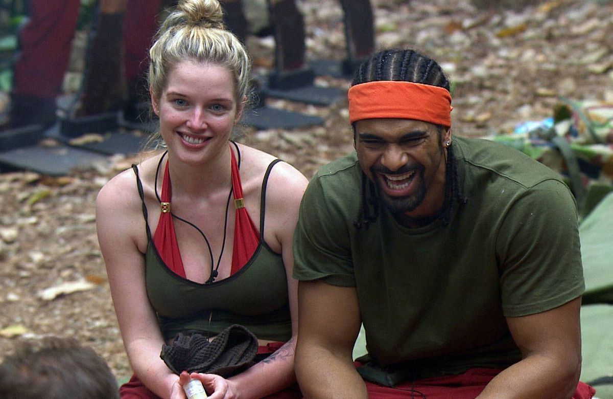Two individuals, one in a green sleeveless top and red shorts, the other in a green tank top and red bikini top, are seated and smiling in an outdoor setting, possibly participating in a survival challenge.