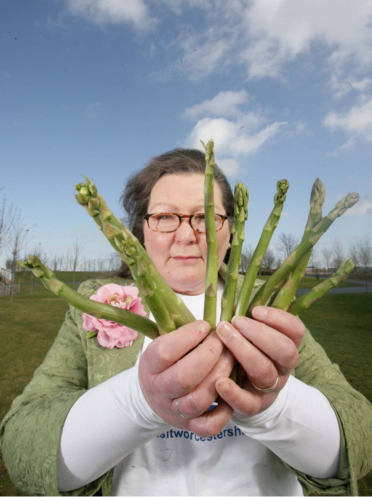 An individual dressed in a green jacket and eyeglasses holds a bundle of green asparagus spears in a field with a blue sky and scattered clouds.