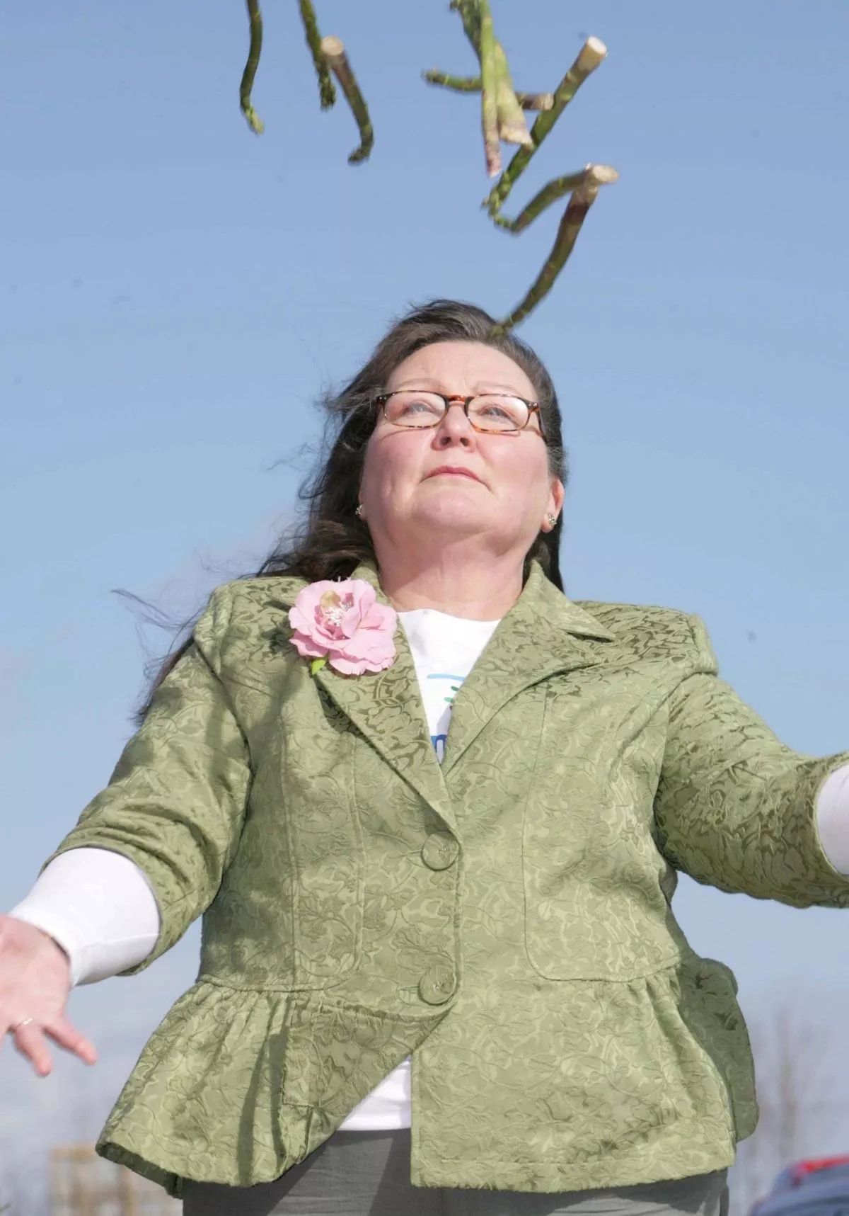 A woman dressed in a green blazer and eyeglasses stands outdoors with her arms outstretched, as if attempting to catch a falling object. The sky is clear and blue in the background.