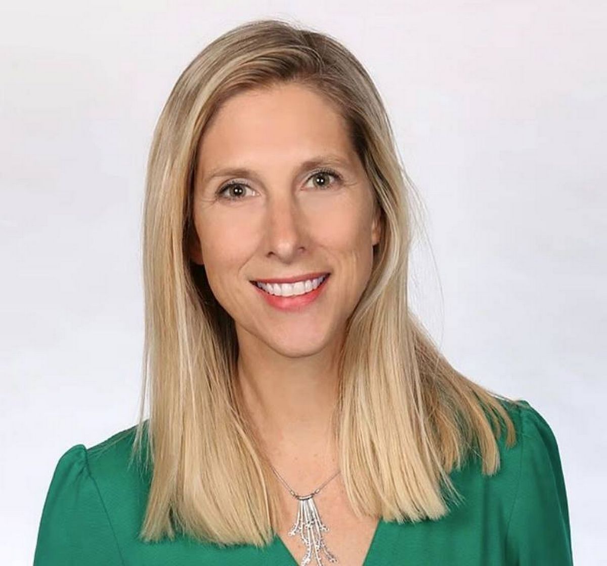 An individual with long, light blonde hair and a green top is seen smiling and looking directly at the camera. The person is positioned against a plain white background, and their expression is cheerful.