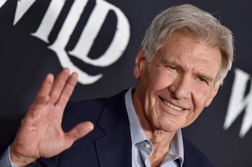 An older man in a suit waves and smiles at a red carpet event