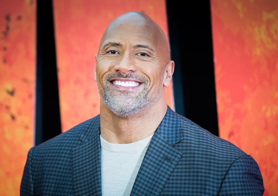 Smiling man in a checkered blazer and light shirt, with close-cropped hair and beard, against a fiery background at a celebrity event