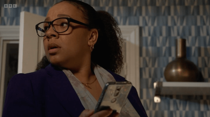 EastEnders Libby Fox Belinda Owusu as Libby Fox holding a phone in EastEnders and looking behind her.