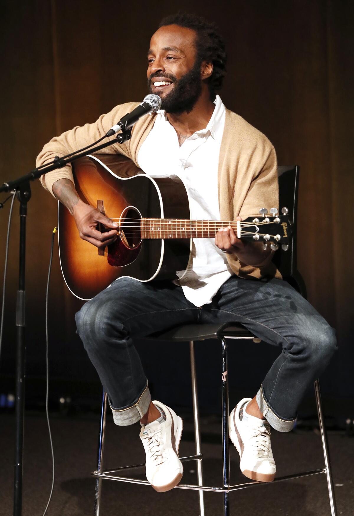 A smiling man sitting on a stool playing an acoustic guitar.