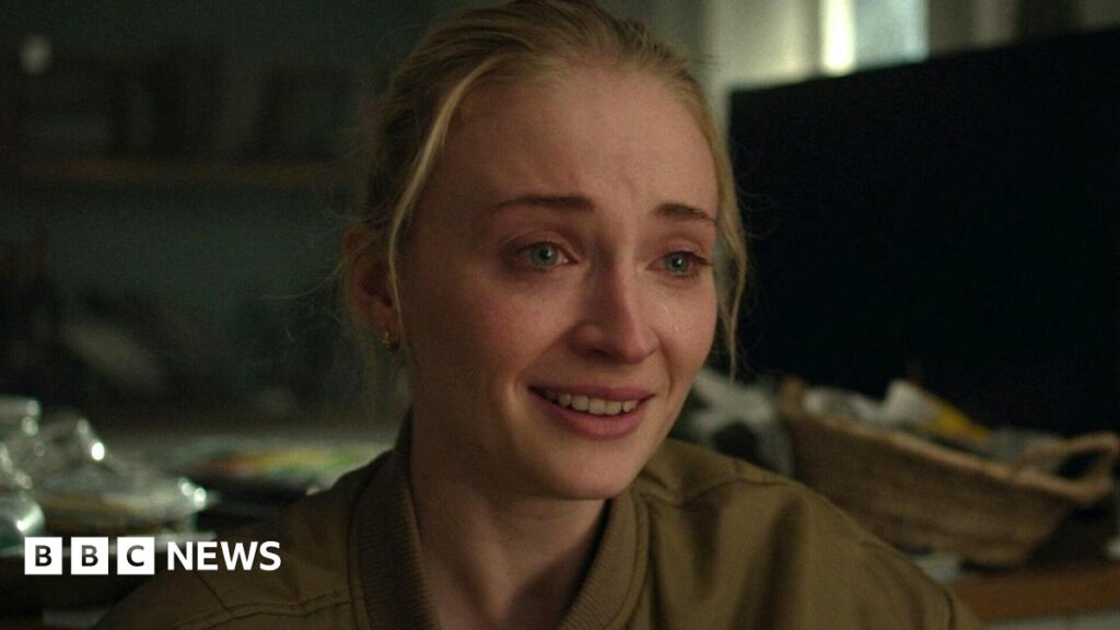 Picture of Sophie Turner looking upset, with tears in her eyes and her hair pulled back. She is wearing a khaki bomber jacket that is slightly unzipped. Messy blurred household items appear in the background.