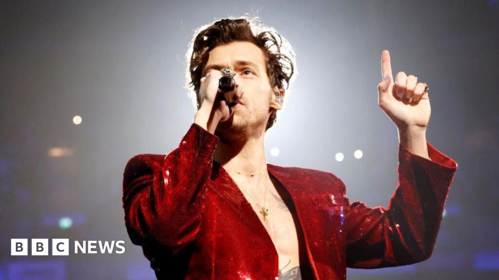 Harry Styles sings into a microphone while performing at the Brit awards in 2023. He has a red, sequinned jacket on and is pointing one finger up towards the ceiling.
