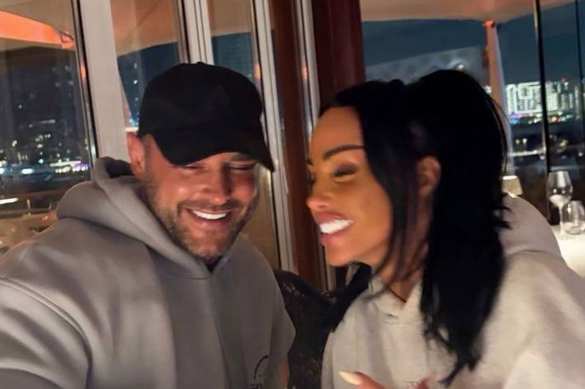 Two individuals, one dressed in a gray hoodie and a black cap, the other in a white hoodie, are smiling and posing for a photograph in an indoor setting with a glass window in the background.