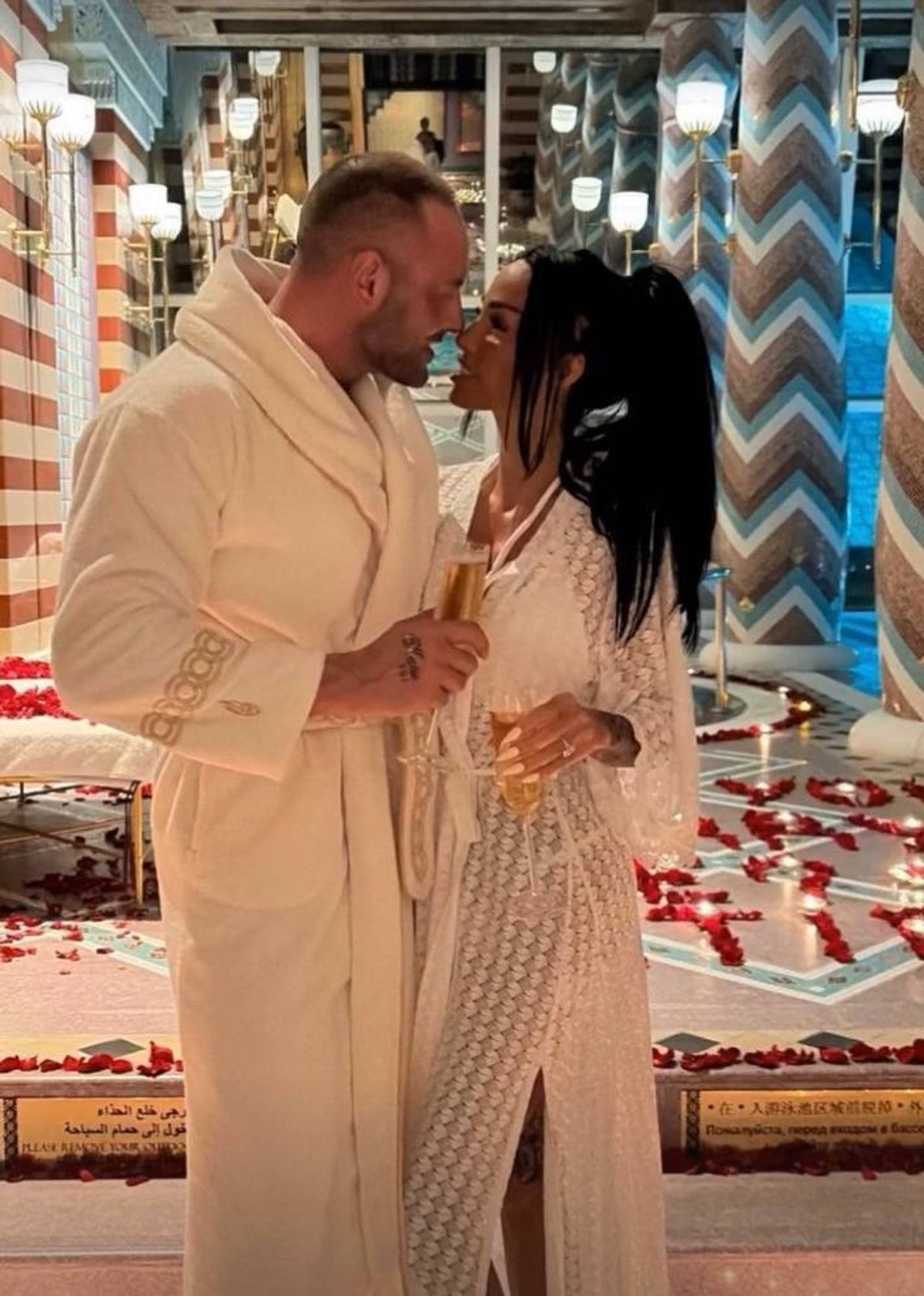 An image depicts a man and a woman standing in a corridor, possibly inside a hotel. They are looking at each other while holding glasses, likely engaged in a romantic moment. The corridor features a distinctive striped and chevron pattern on the walls.