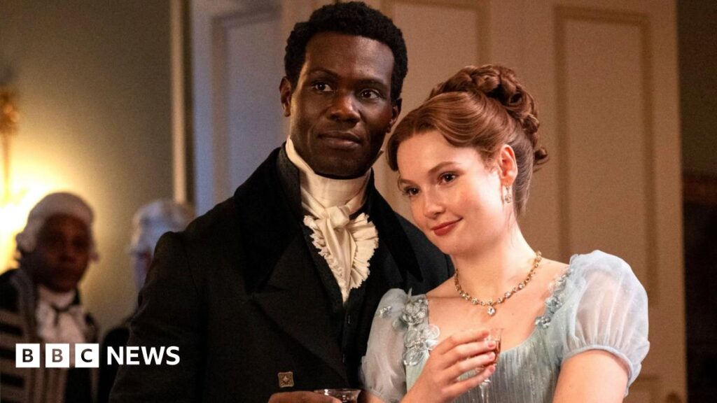 Francesca (Hannah Dodd) and John (Victor Alli) survey the scene at yet another social gathering. John is wearing a black jacket and white shirt, while Francesca is in a light blue dress with short chiffon sleeves.