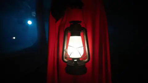BBC A red-cloaked figure carrying a lamp on The Traitors