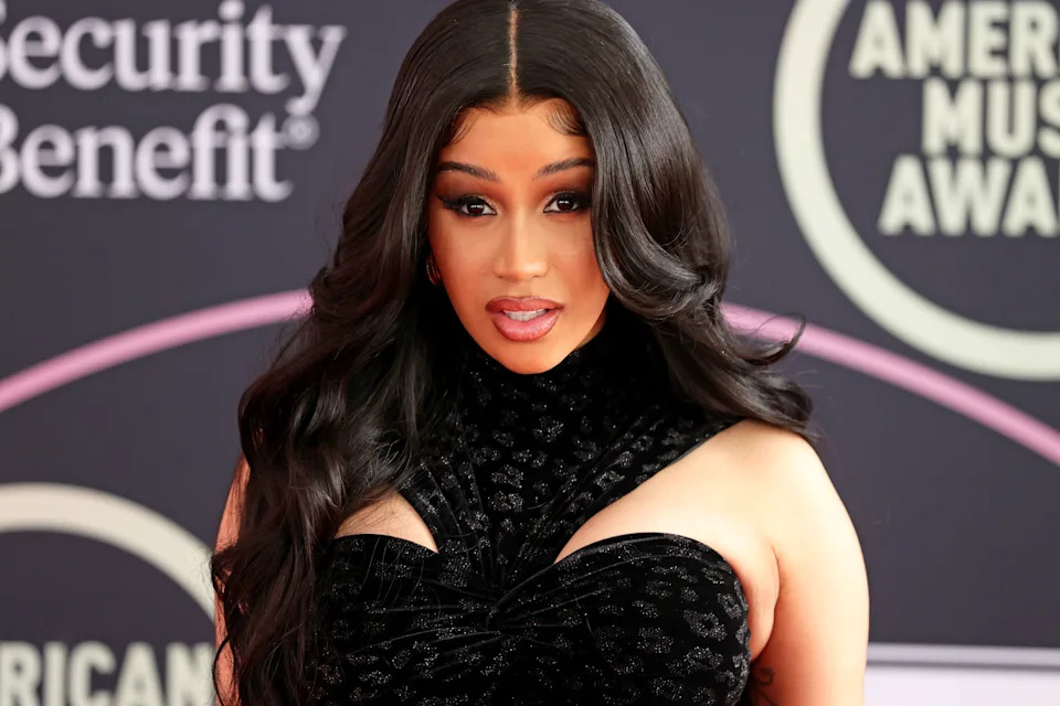 A person in glamorous evening attire poses on a red carpet at the American Music Awards, with long wavy hair and an elegant dress