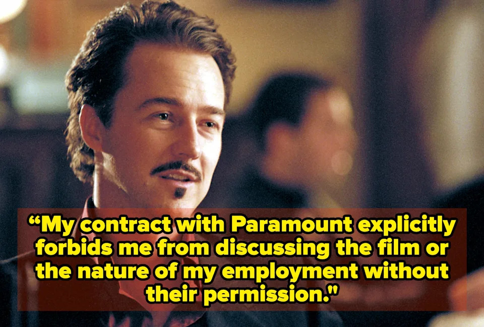 A person with short hair and a mustache is sitting in a relaxed setting, wearing a dark suit and an open-collared shirt, looking off to the side