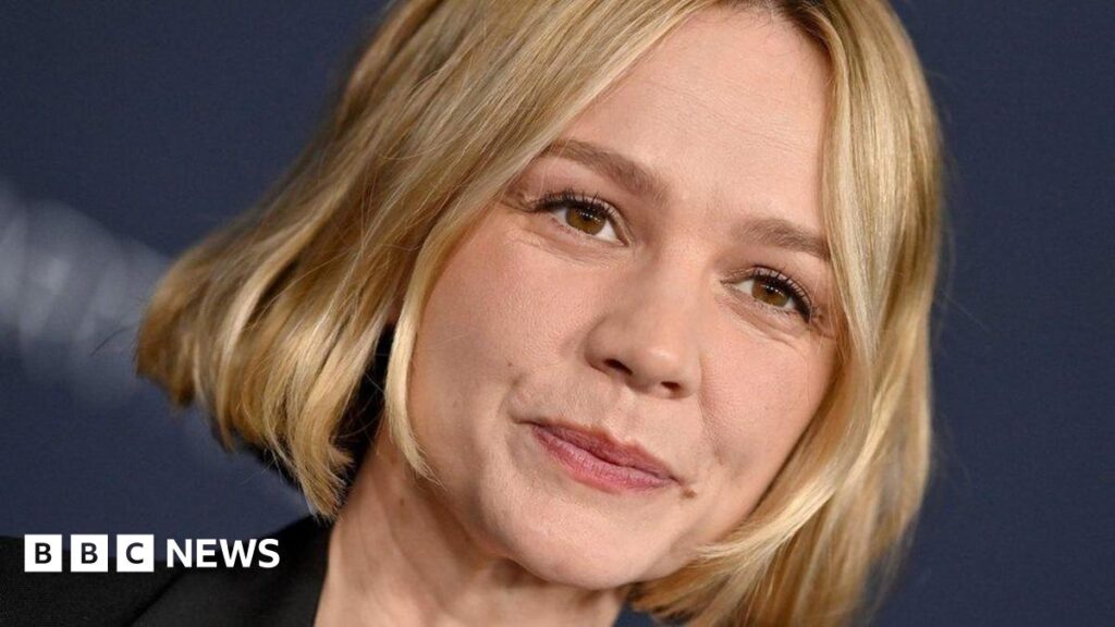 Carey Mulligan smiling at the camera, she has short blonde hair, brown eyes and a black jacket, with a navy blue background.