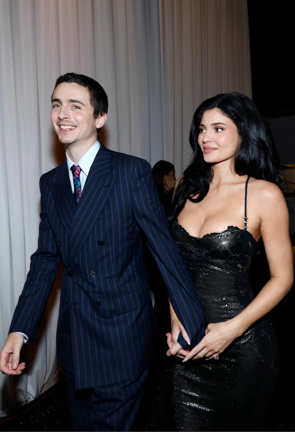Timothee Chalamet holding hands with Kylie Jenner at the Critics Choice Awards. He is wearing a dark navy pinstriped suit and she is in a black gown.