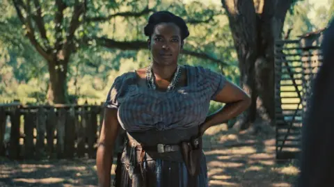 Warner Bros. Pictures Wunmi Mosaku, who has her left hand on her hip and is staring off camera, is pictured as Annie in Sinners. 