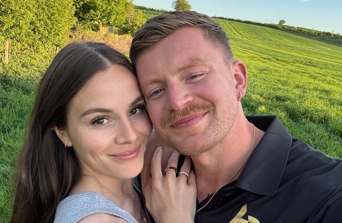 A man and a woman are standing close together, smiling and posing for a photograph in an outdoor setting with a lush green field in the background.