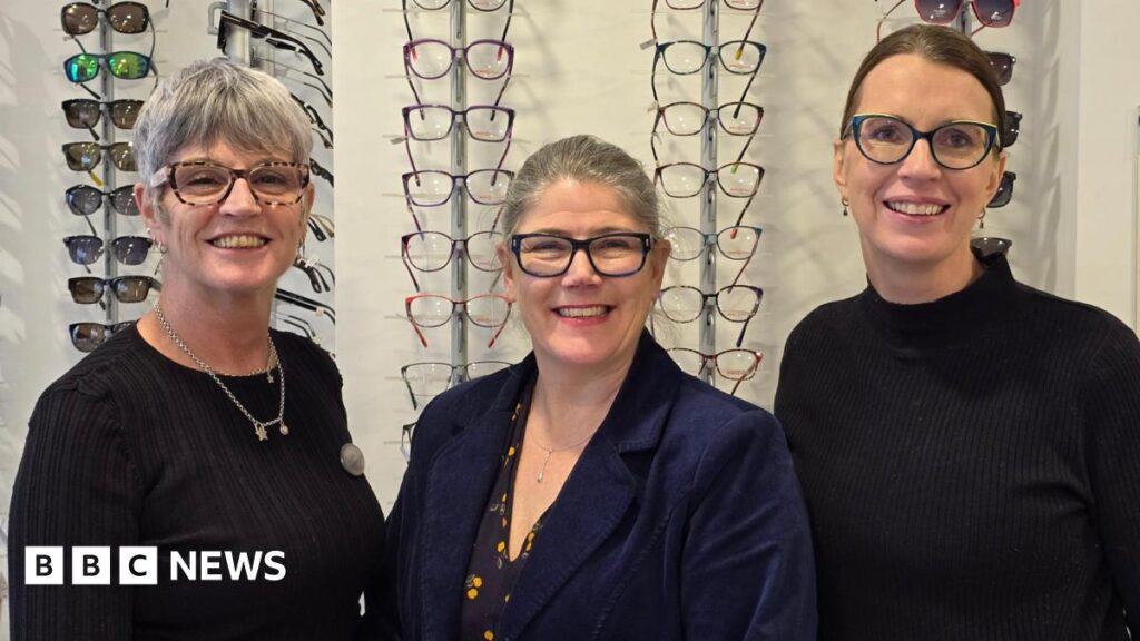Three women who are all wearing glasses smile at the camera. They are standing in front of display racks of spectacles, with a rack of sunglasses to the left and right.