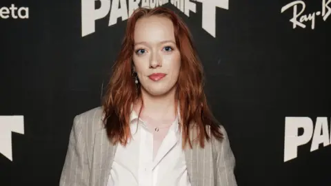 Darren Gerrish/Getty Images Marybeth is standing in front of a black backdrop at a film premier. She has ginger shoulder-length hair and is wearing a white shirt and grey pin stripe suit. 