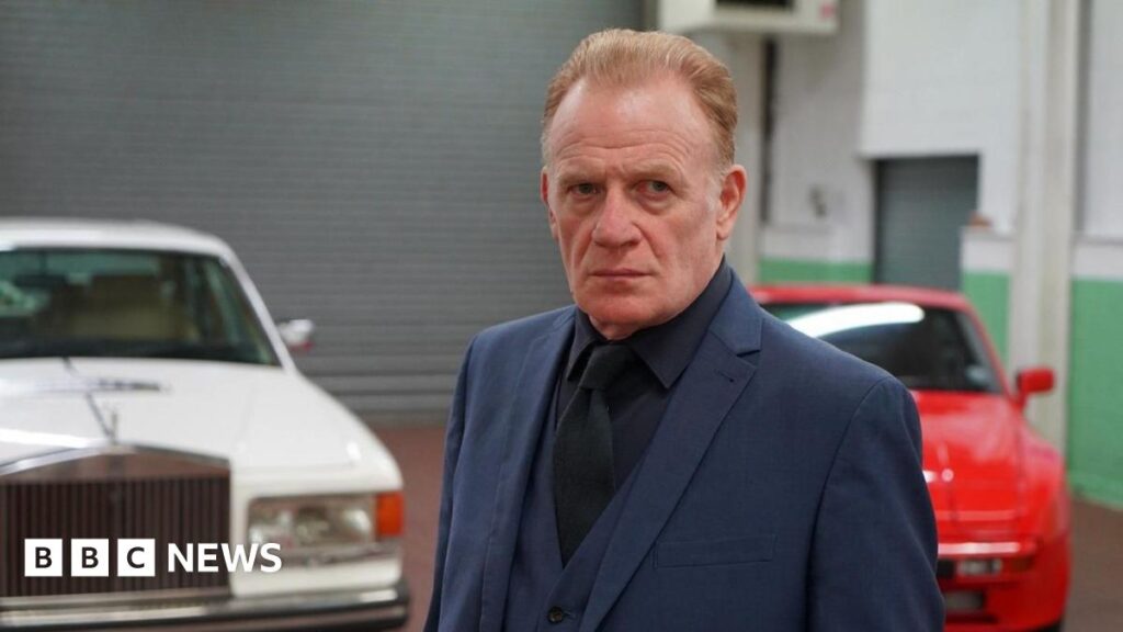 Netflix Welsh-language show Dal y Mellt hopes to build on success Mark Lewis Jones, wearing a blue two piece suit with a dark shirt. He has light brown/ginger hair. Pictured behind him are two cars; one white and one red.