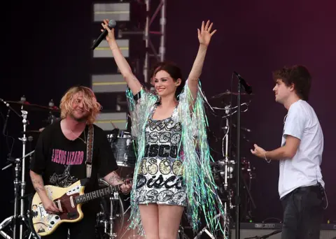 Getty Images Sophie Ellis Bextor on stage with Royel Otis. She has her arms raised to the sky, in a sparkly dress with the word "disco" stitched onto it in a variety of fonts. The band smile as they look at her.