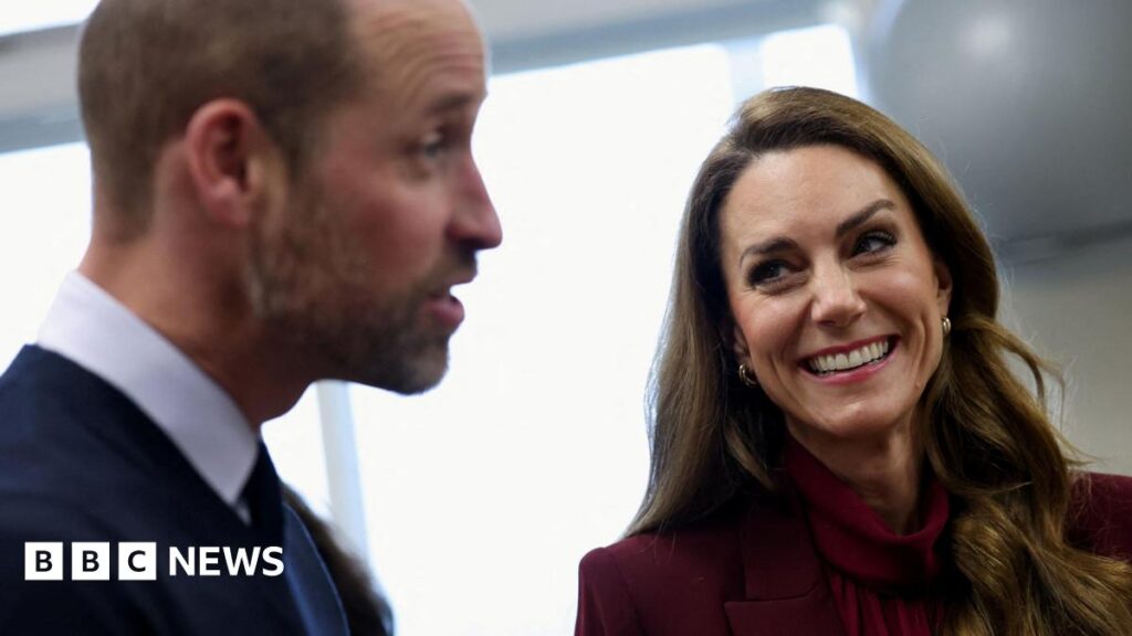 The Prince and Princess of Wales, joint Patrons of NHS Charities Together, during a visit to Charing Cross Hospital, west London. William is pictured to the left of the image and is looking to the right, with his eyebrows raised. Catherine is on the right, smiling at him. Her long brown hair is down and she is wearing a burgundy trouser suit.