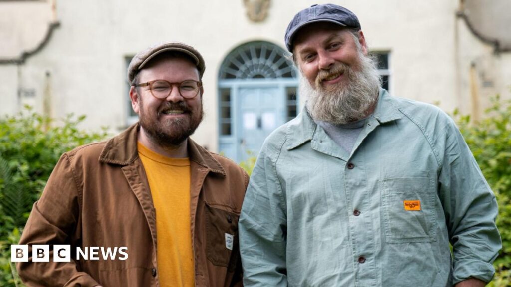 Banjo Beale’s latest project isn’t close to home – it is his home Banjo Beale and his husband Ro stand in front of the derelict mansion Ulva House, a grand but shabby building - its pale blue front door visible behind them. Banjo, with his trademark beard, glasses and flat cap, smiles alongside Ro, also in a cap, who has a greying long beard.