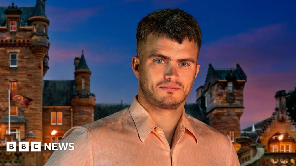 Traitors contestant Matthew Hyndman, with short brown hair and stubble, wearing a light pink shirt. He is standing in front of the Traitors castle and it is nighttime.
