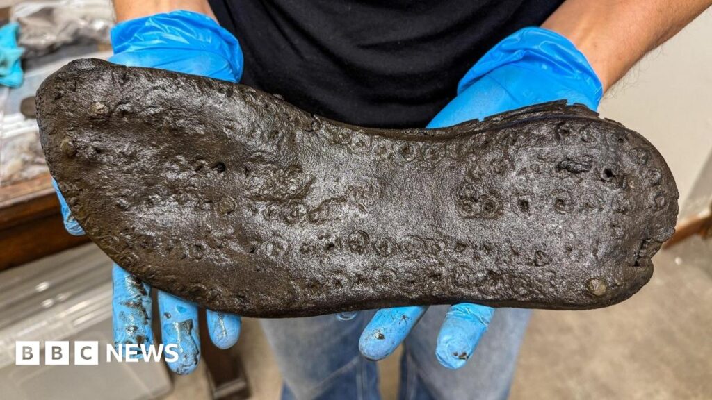 Northumberland fort’s large Roman shoes to go on display Dr Elizabeth Greene wearing a black T-shirt and blue gloves with sunglasses hanging from her top. In her hands are the sole of a large black shoe.