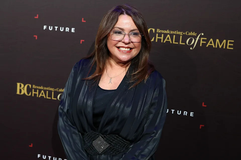 Person smiling on the red carpet, wearing a dark, elegant outfit with glasses, attending a Hall of Fame event