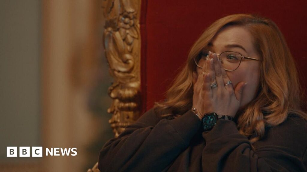 Sarah Taylor, a woman in her thirties, sits on a throne-like chair with her hands covering her face in disbelief.