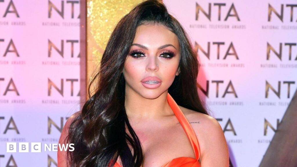 Jesy Nelson in an orange dress posing in front of an NTA-branded backdrop at the 2020 National Television Awards