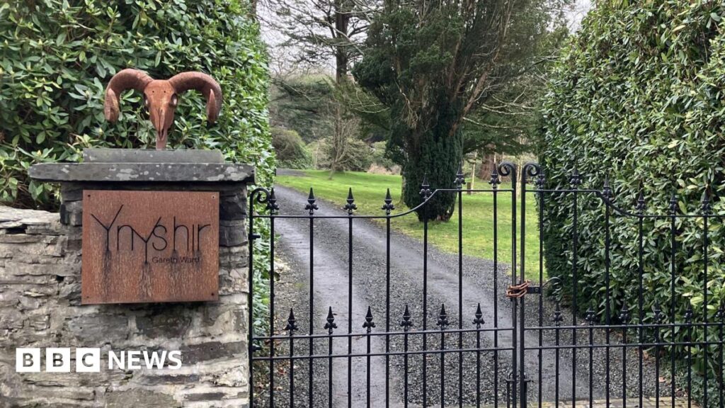 The gated entrance to Ynshir restaurant. A black cast iron gate leads up a drive. On the wall is a bronze coloured sign with the restaurant's name on it.