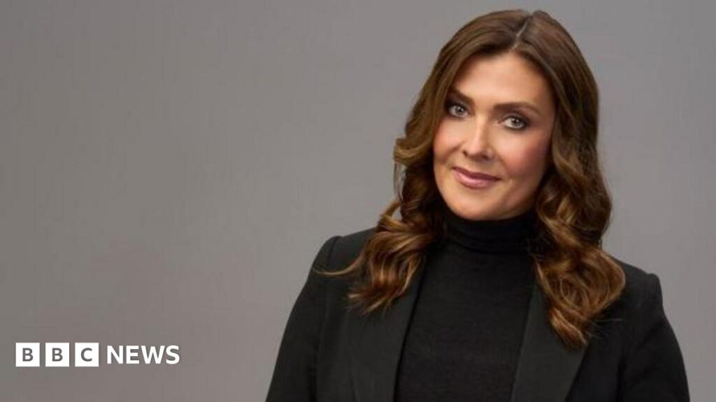 Kym Marsh says she has ‘big shoes to fill in Single White Female’ Kym Marsh, who is wearing a black blazer and black polo neck jumper, is standing in front of plain grey background for this posed photograph and is smiling.