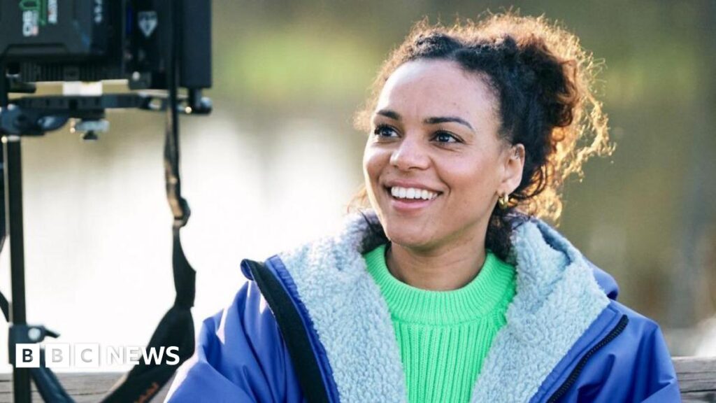 Swim Sistas to screen at Northampton Film Festival Cat White, a filmmaker, sitting down looking into a camera monitor. She is wearing a bright green jumper with a blue fleece-lined coat over the top. She is smiling but not looking at the camera.