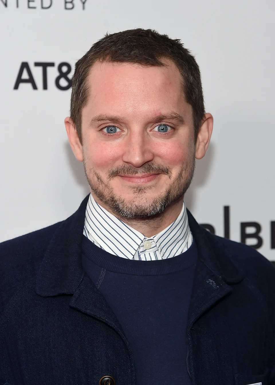 A person smiling at a media event, wearing a striped shirt and dark jacket