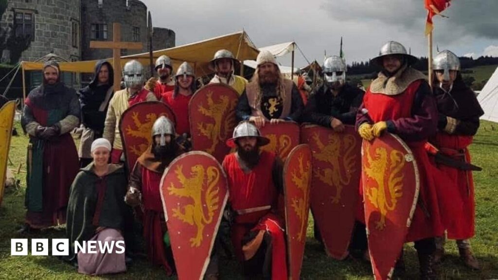 12 men and one woman in medieval costume. The men are in red soldier uniforms and the woman is wearing a pink skirt and green cloak. They are standing on a field