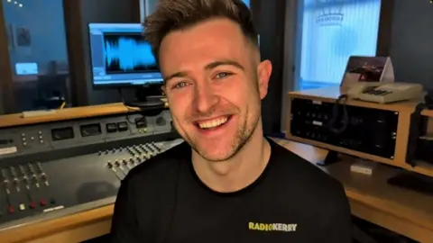 Brendan Fuller smiling at the camera in a radio studio. He has short, spikey blonde hair and is wearing a black t-shirt with a "Radio Kerry" logo on the chest.