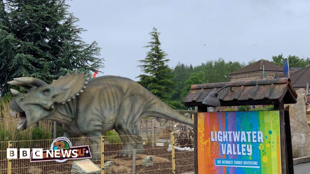 A large model of a Triceratops dinosaur stands within a fenced area at Lightwater Valley amusement park. Behind it, a colourful sign reads "Jurassic Adventure Golf". Another vibrant sign proclaims "LIGHTWATER VALLEY – THE ULTIMATE ADVENTURE".