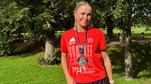 Leah Walsh Leah smiling looking at the camera. She is stood in front of trees and greenery and wears a red Cardiff half marathon finishers t-shirt, a black medal and black running shorts. Leah has blonde hair in a high ponytail. 