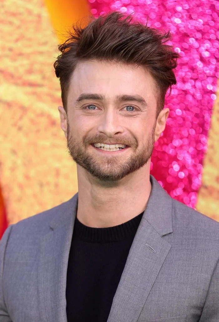 A person on a red carpet in a gray blazer and black top, smiling with a glittery backdrop