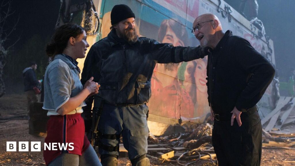 Stranger Things documentary-maker gives reason for last minute script Millie Bobby Brown and David Harbour in character as Eleven and Jim Hopper. Eleven wears red shorts over grey leggings and a grey jumper. Jim Hopper wears a black leather jacket, a black beanie hat and dark trousers.They are speaking to director Frank Darabont on the set of Stranger Things. There is a vintage advertisement posted in the background with debrief on the ground.