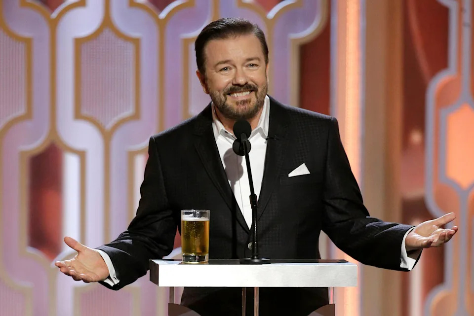 A person stands on stage behind a podium with a drink, smiling and speaking into a microphone. They are wearing a dark suit with a white pocket square